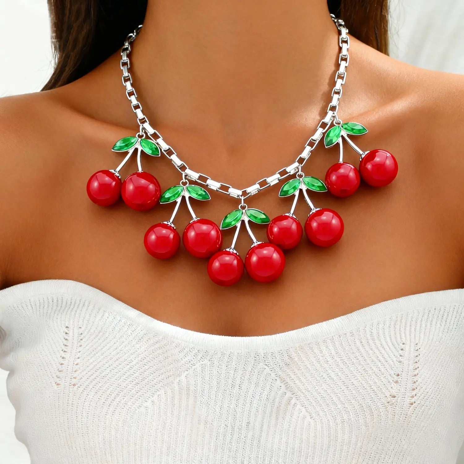 Un collier statement de cerises rouges, avec des cerises rouges brillantes en résine et des feuilles émaillées vertes suspendues à une chaîne de collier ras du cou en cristal à tonalité argentée.