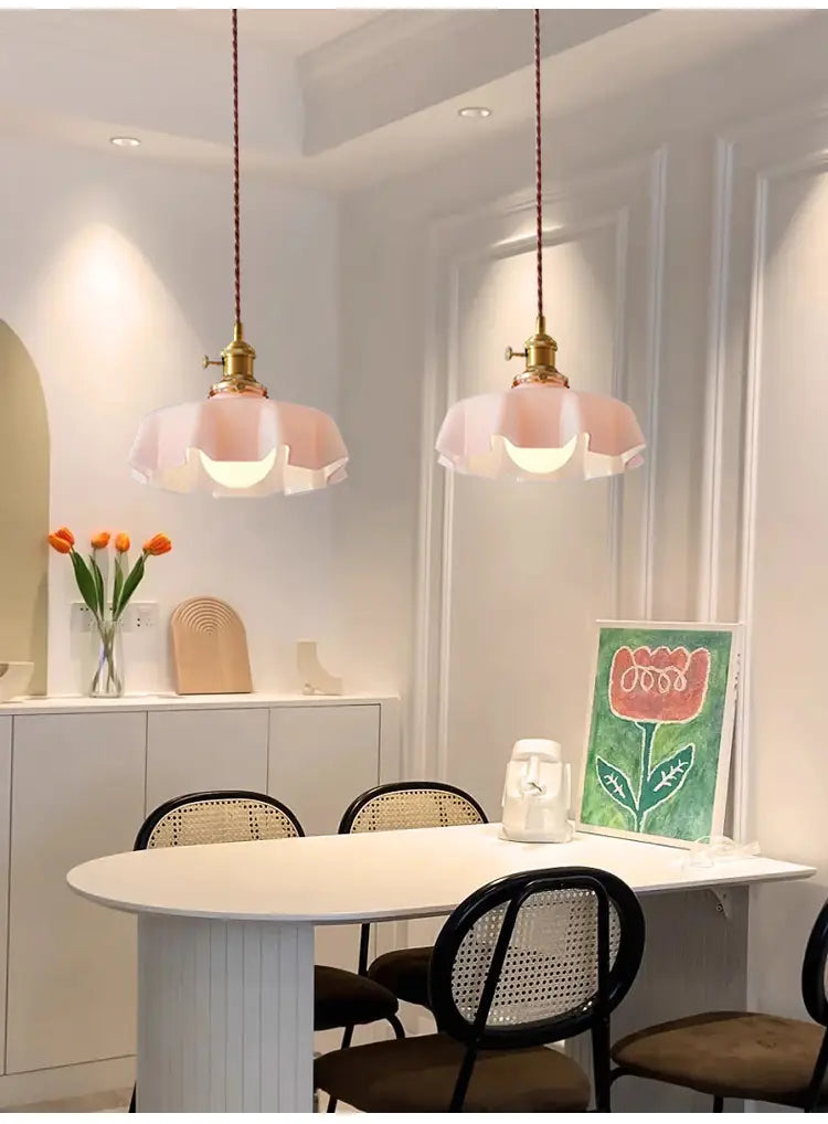 Deux luminaires suspendus avec des abat-jours en verre à jupe florale rose et des têtes de lampe en cuivre, suspendus à des cordons tressés au-dessus d’une table à manger blanche.