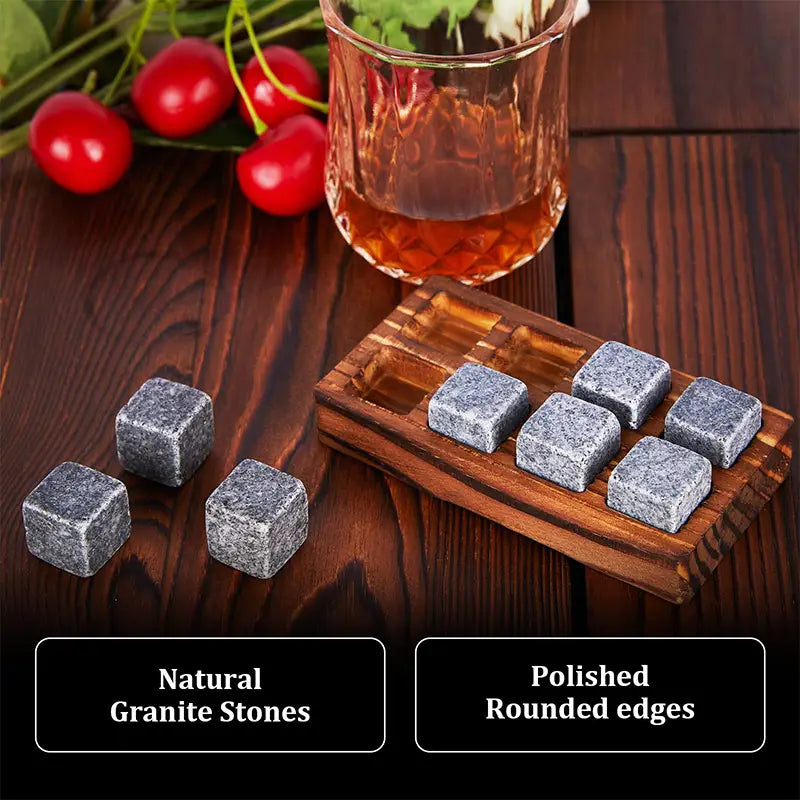 Set aus natürlichen grauen Granit-Whiskey-Steinen mit polierten abgerundeten Kanten, präsentiert auf einem hölzernen Tablett neben einem Glas bernsteinfarbener Flüssigkeit und Kirschen in einem Whiskey-Steine-Geschenkset mit Gläsern und Holzbox.