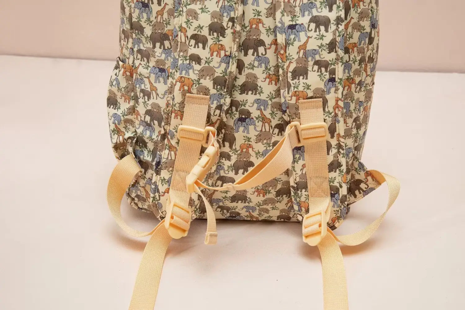 Sac à dos dinosaure pour enfants de couleur beige, avec un motif ludique d’éléphants et de girafes aux tons terreux atténués, et des bretelles réglables jaune clair, pour un usage scolaire.
