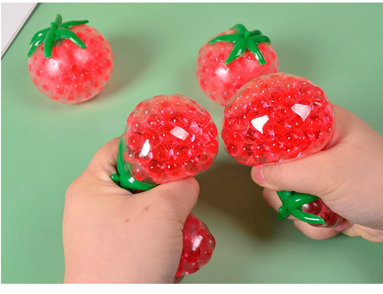 Red strawberry-shaped squishy tomato stress relief toy with green leafy top and translucent gel beads inside for squeezing and stress relief.