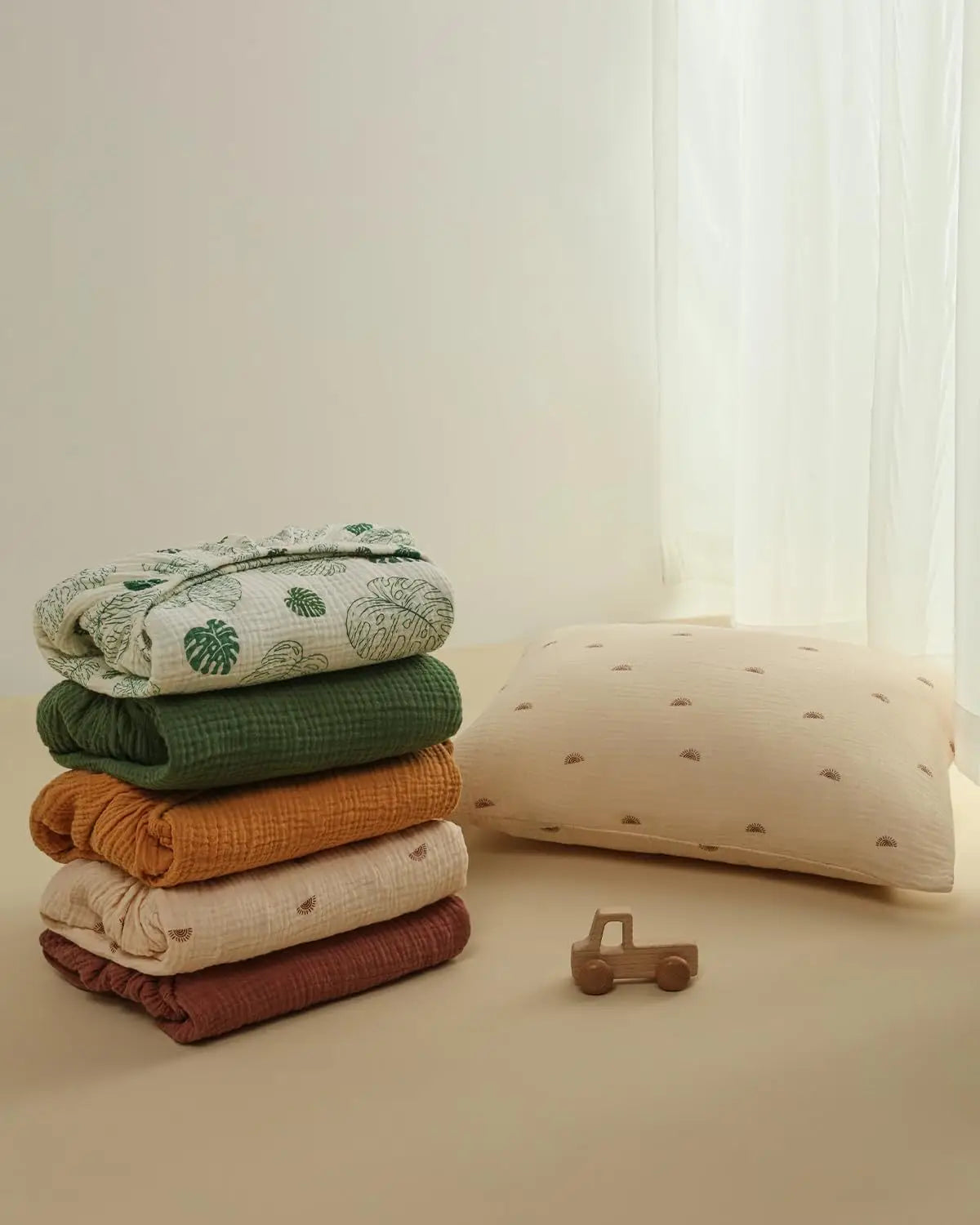 ein Stapel gefalteter Babybettlaken-Set-Decken aus Baumwollmuslin in erdigen Farbtönen mit dezenten Blatt- und Punktmustern.