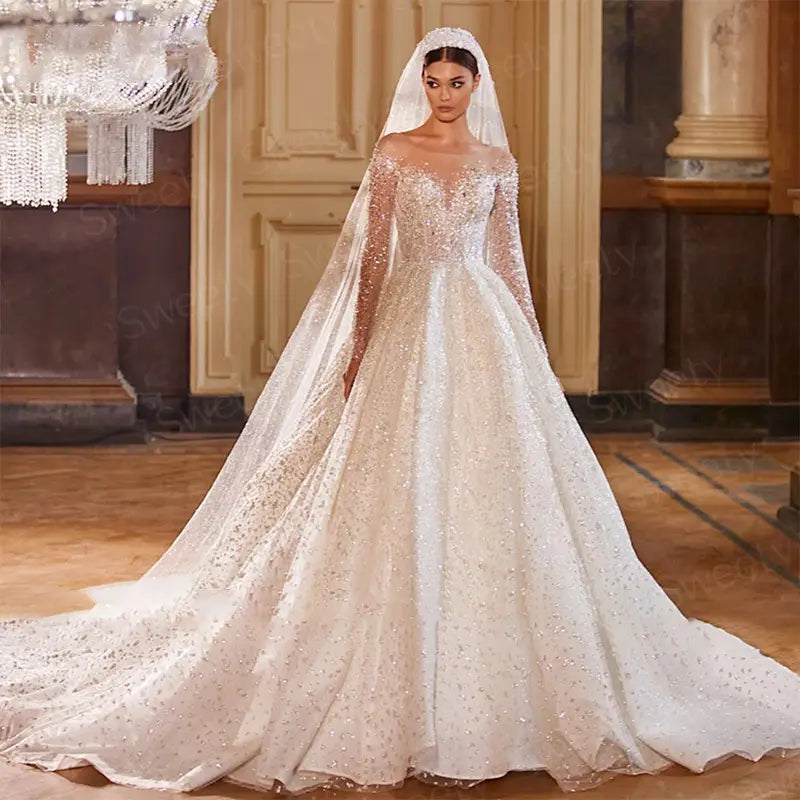 Une robe de mariée en style boule ivoire étincelante, perlée, avec de longues manches transparentes et une traîne fluide, rehaussée d’un empiècement de perles minutieusement travaillé.