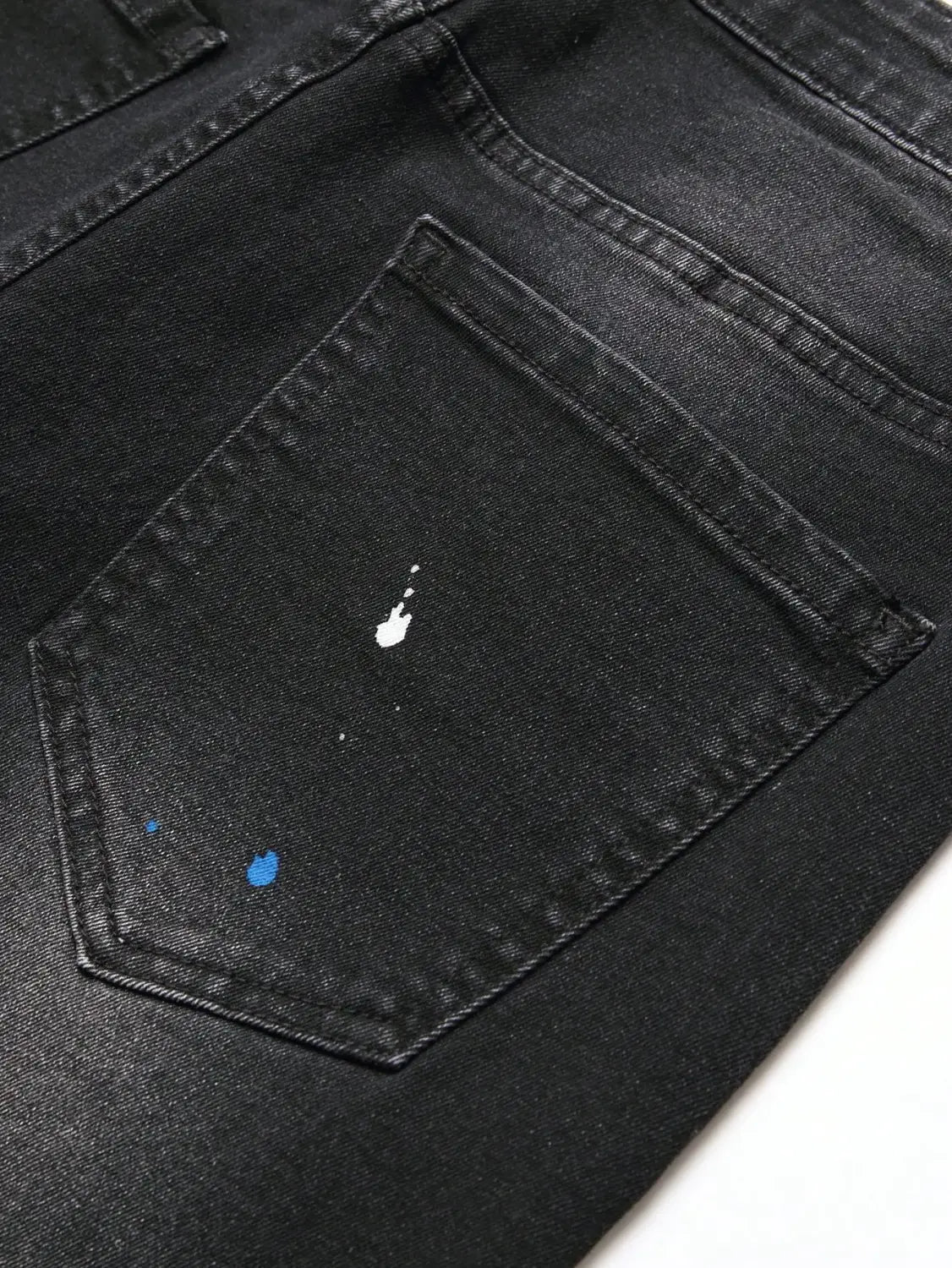 Face arrière des jeans denim skinny extensibles déchirés Y2K pour hommes en noir, présentant un motif discret d’éclaboussures de peinture blanc et bleu sur la poche arrière, réalisée dans un matériau denim extensible.
