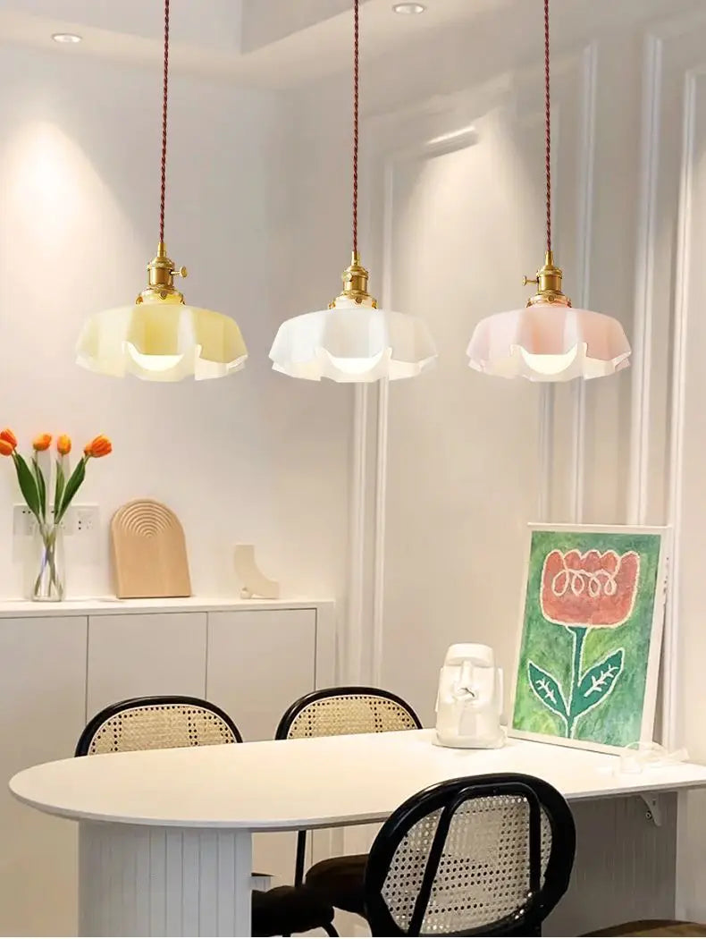 Trois luminaires suspendus à jupe florale en verre avec têtes de lampe en cuivre et abat-jours en forme de pétales en tons jaune clair, blanc et rose, suspendus au-dessus d’une table à manger.