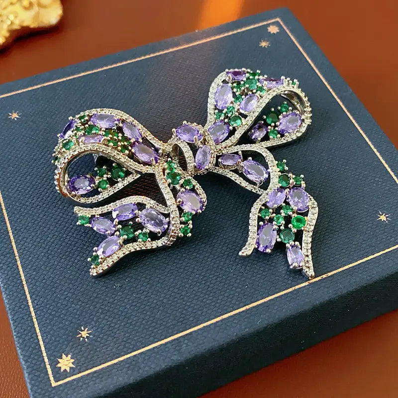 Eine funkelnde silberfarbene Zirkon-Schleifenbrosche, geschmückt mit oval geschliffenen lila Amethysten und smaragdgrünen Edelsteinen, die luxuriöses französisches Schmuckdesign ausstrahlt.