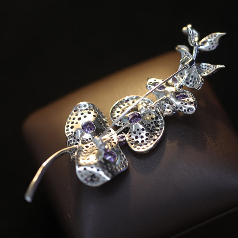 Broche de orquídea en tono de plata que presenta pétalos perforados con gran detalle y gemas violetas incrustadas en un pin de lujo de diseñador de mariposa de zirconio.