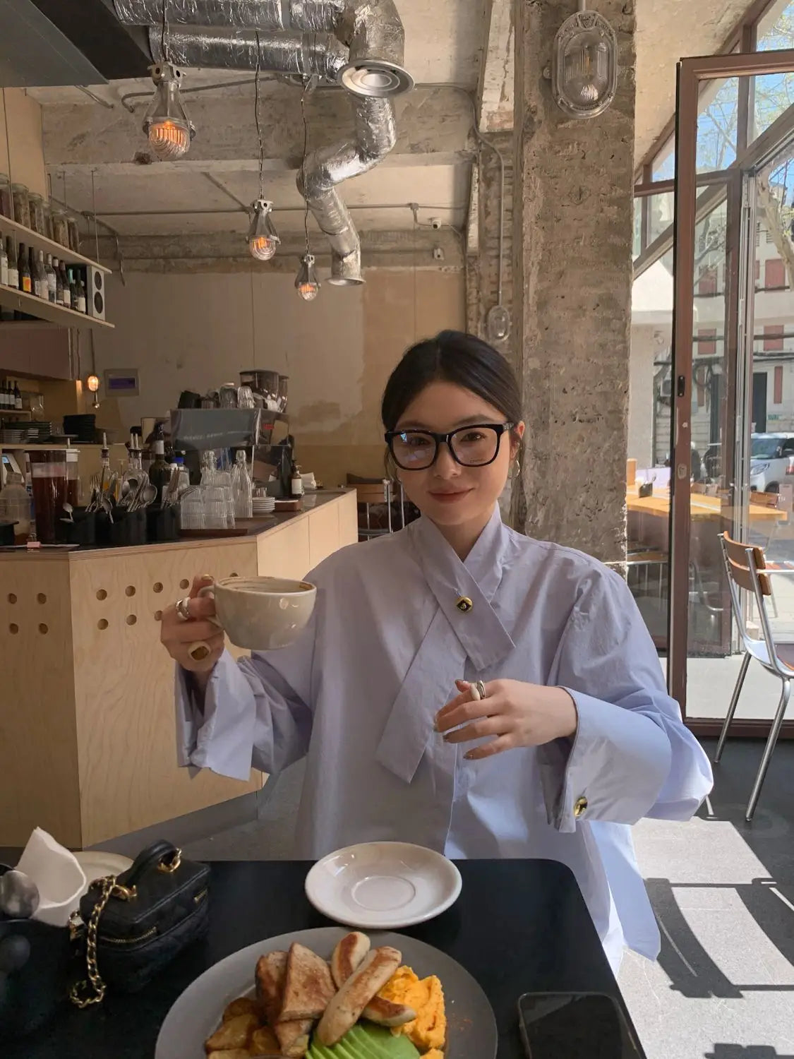 A woman wearing a light blue oversized button up shirt with long sleeves and gold buttons holds a beige ceramic mug.
