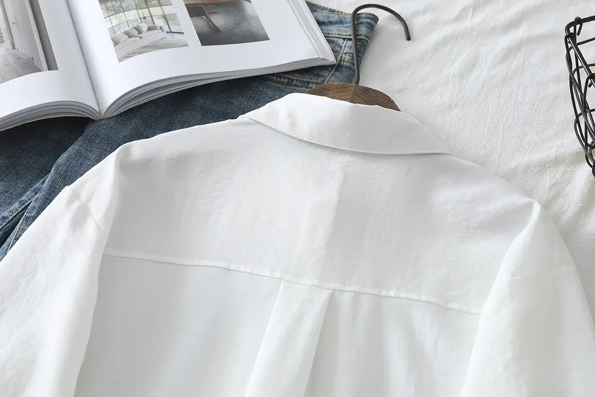 Eine knusprig-weiße Damen-Casualbluse aus Baumwollmischgewebe mit langen Ärmeln mit klassischem Kragen und sauberer Verarbeitung, auf einem Holzkleiderbügel präsentiert.