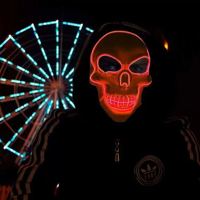 Un disfraz de calavera de Halloween con máscara luminosa, que presenta una máscara de calavera de neón roja luminosa con cuencas de ojos negras y una boca sonriente, colocada sobre una sudadera oscura con paquete de controlador para efectos de luz fría
