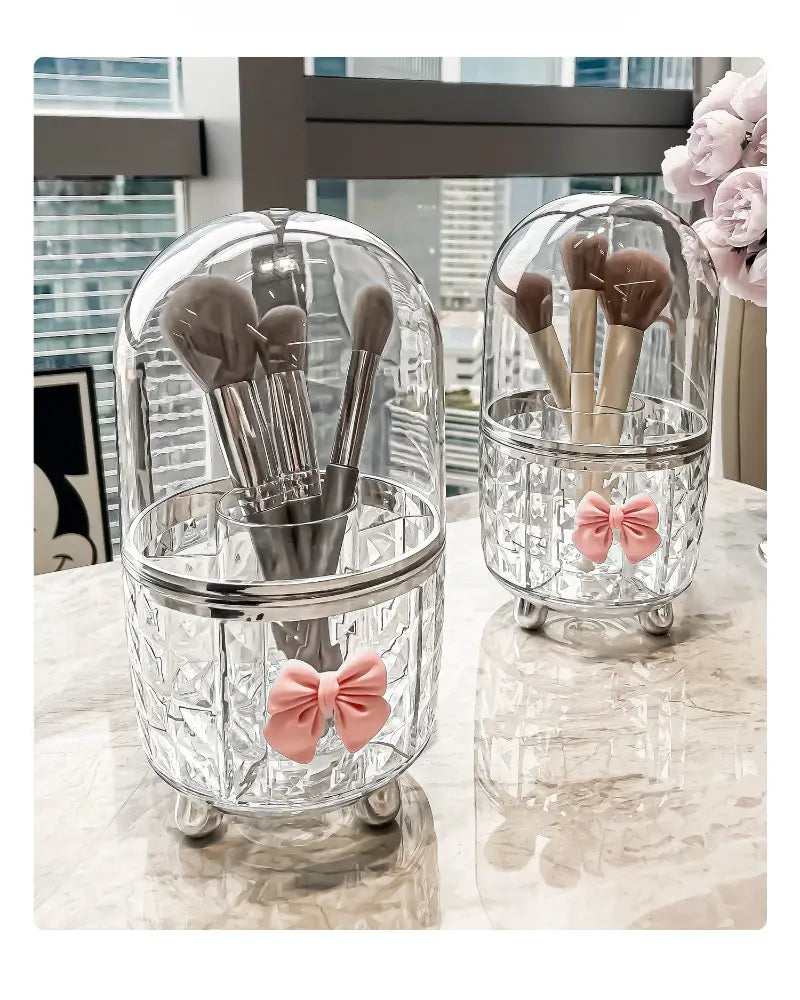 Porte-pinceaux en verre transparent en forme de dôme, avec des accents argentés et des décorations de nœud rose, dans une boîte de rangement tournante pour maquillage et organisation des cosmétiques pour un usage de rangement.