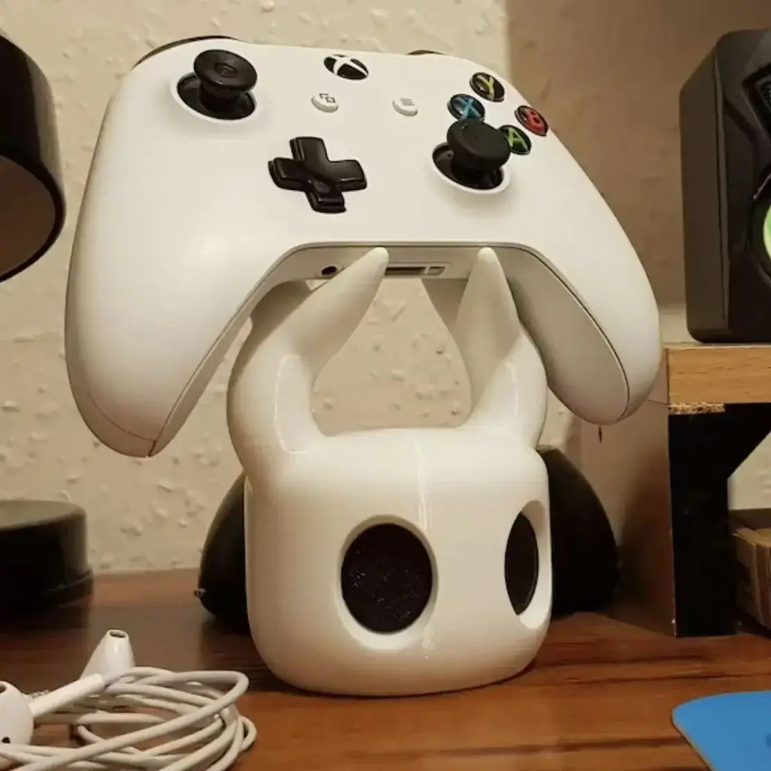 White Xbox controller resting on a white hollow knight game controller stand for PS5 featuring a skull-shaped design with black eye holes and horn-like protrusions.