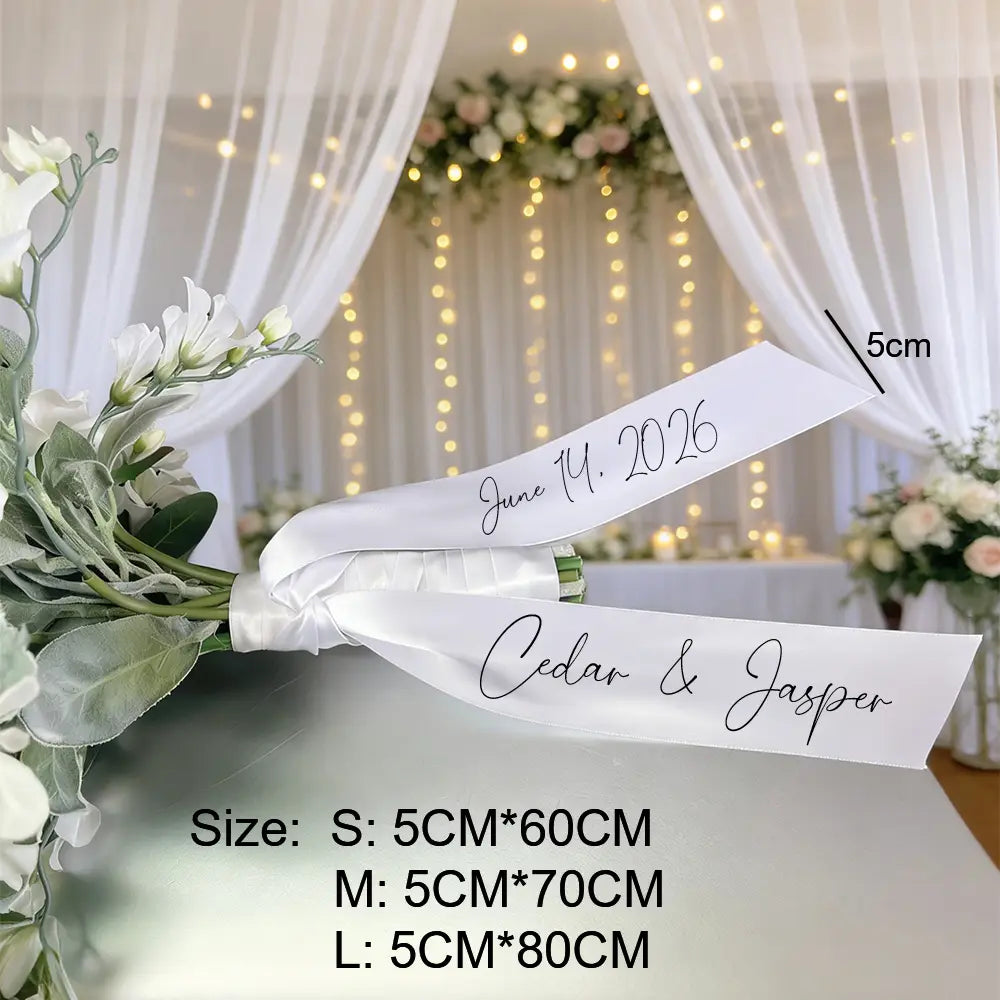 Un ruban de bouquet de mariage en satin blanc personnalisé avec un lettrage cursif noir affichant les noms propres cedar et jasper ainsi que la date 14 juin 2026, noué autour d’un bouquet de fleurs blanches.