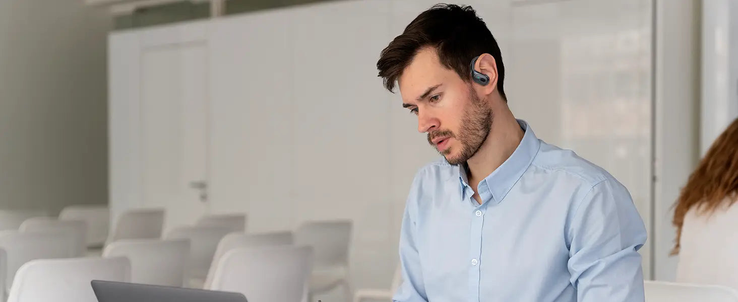 Un homme portant des écouteurs sans fil bleus avec des crochets d’oreille dans un environnement de bureau, avec un boîtier de charge pour la recharge des écouteurs sans fil.