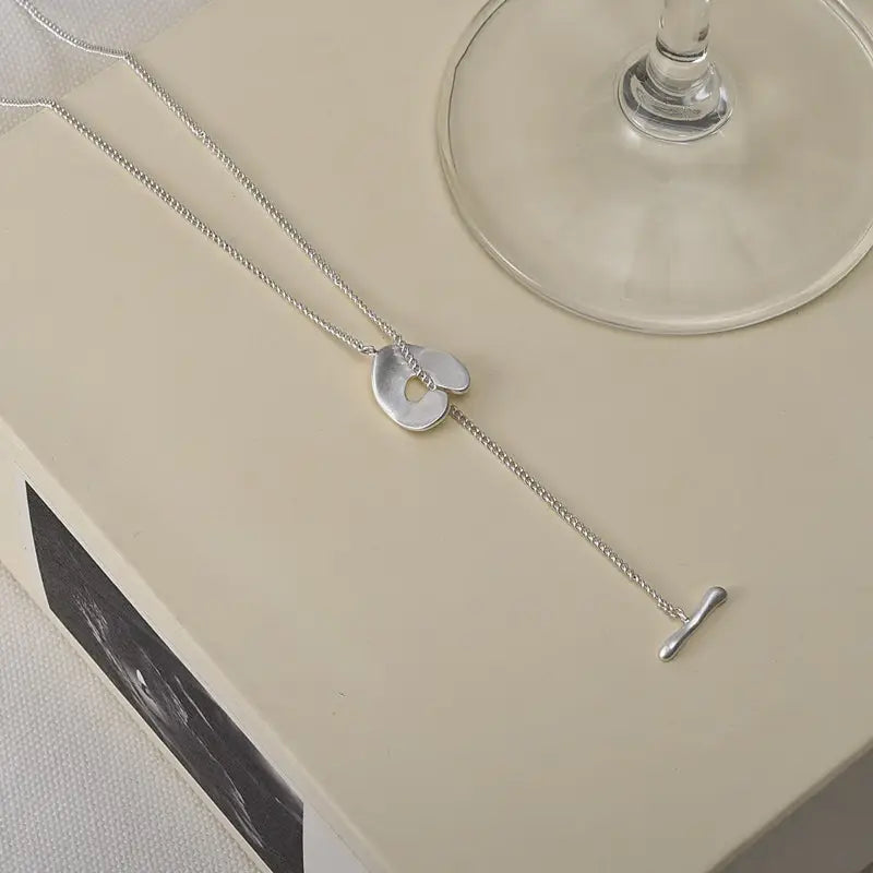 Collier délicat à chaîne en argent pour femmes, présentant un pendentif abstrait en forme de cœur lisse, de forme abstraite, avec un trou central, et une petite attache à t-bar, dans un design de collier pendentif à superposition géométrique doré.