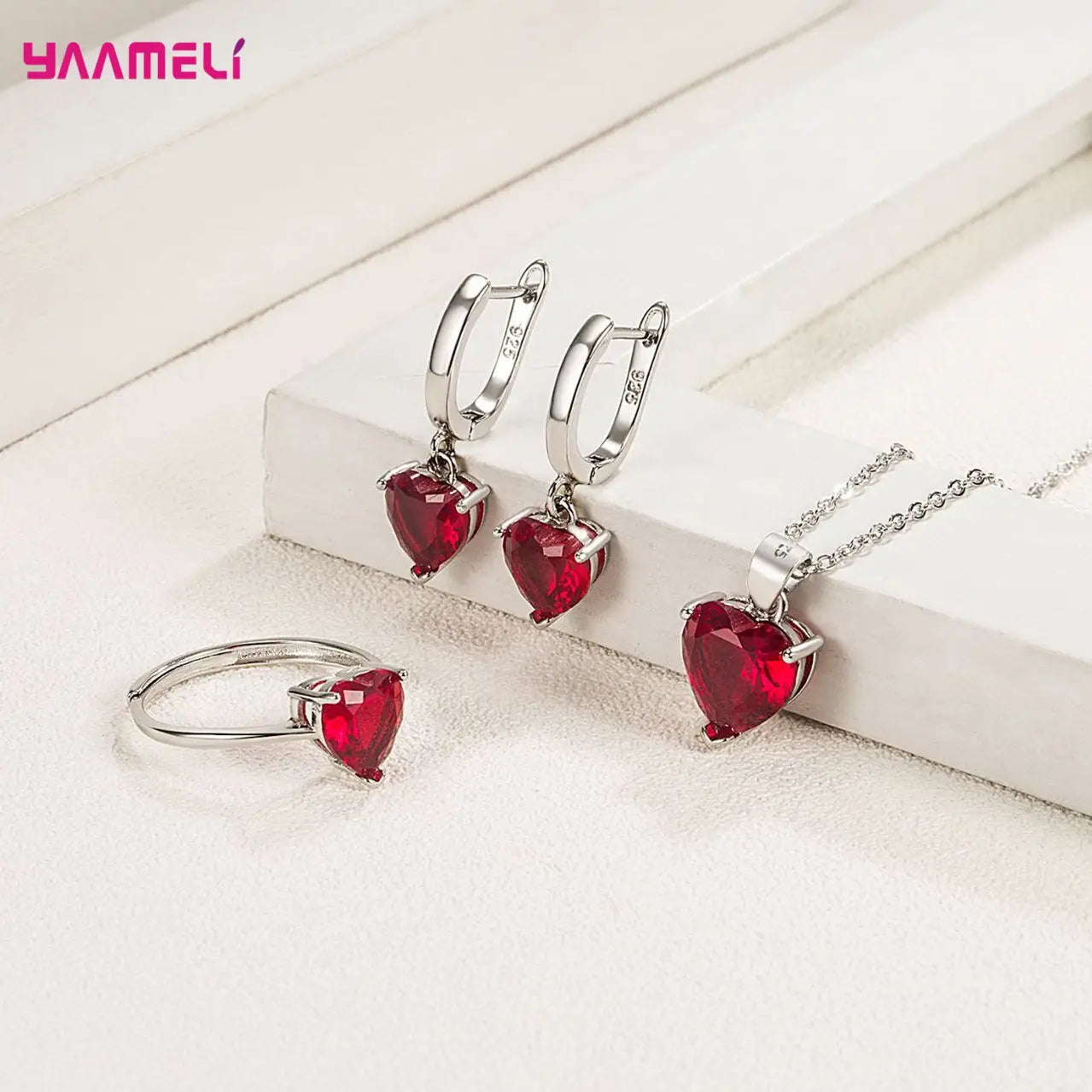 A set of sterling silver heart-shaped jewelry featuring vibrant red zircon gemstones including earrings huggies necklace and ring.