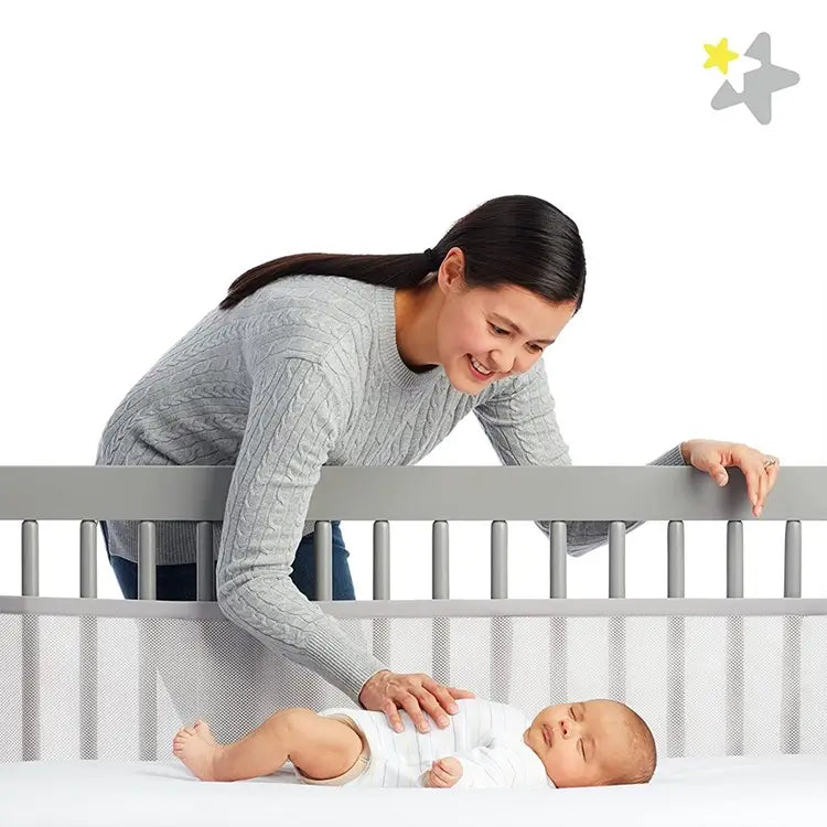 Graues Babybett mit atmungsaktiven Mesh-Seiten und vertikalen Lamellen, mit Teddy-Bären-Bettwäsche und einem Mesh-Krippenstoßschutz.