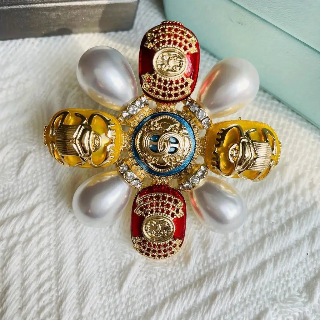 Broche de perles designer en forme d’insecte coléoptère, épingle sertie de strass, présentant un emblème central vibrant bleu et or, entouré de quatre scarabées dorés aux accents d’émail rouge, avec des perles blanches.