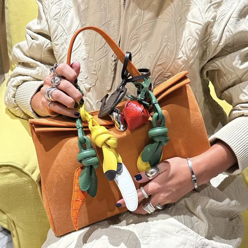 Un sac à main en daim orange orné de charms en cuir végétal aux thèmes colorés et d’accents métalliques, conçu comme ornements de sac à main.
