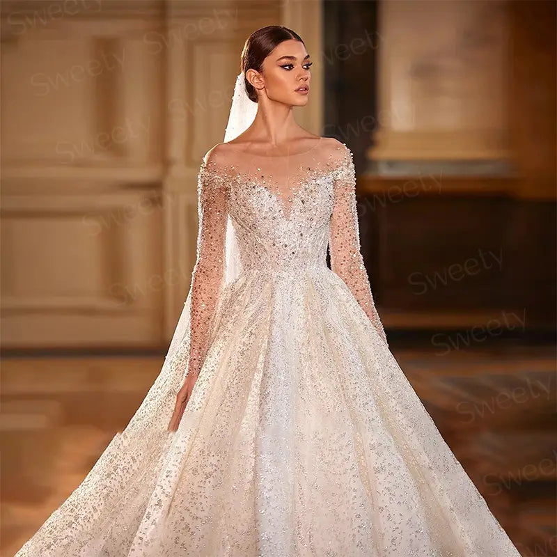 Une somptueuse robe de mariée en dentelle ivoire perlée, de style boule, avec un décolleté en épaules dénudées et de longues manches transparentes.