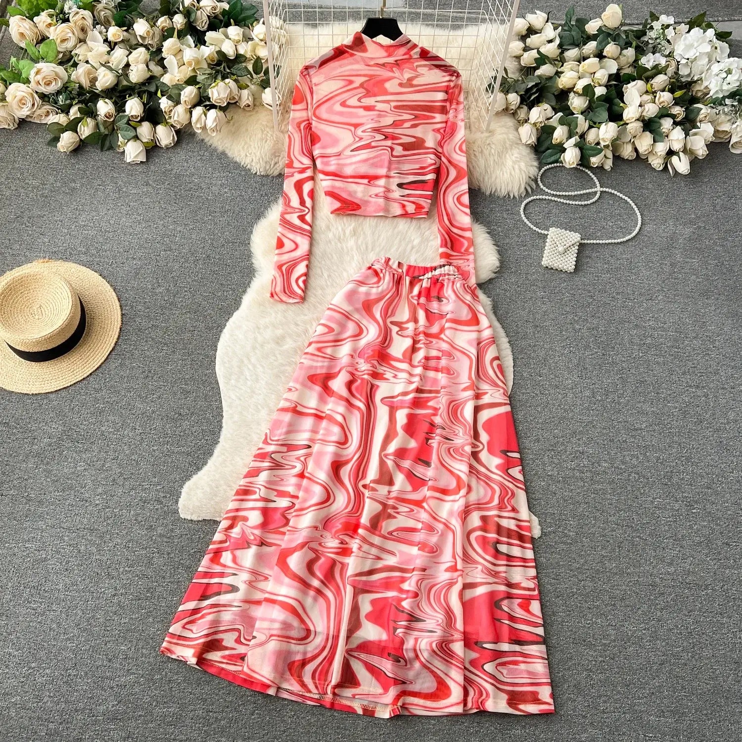 A vibrant pink and red marbled two piece mesh skirt set featuring a high neck cropped top with long sleeves and a flowing satin like maxi skirt with elastic closure.