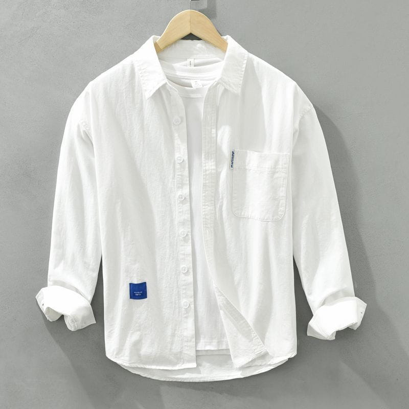Camisa de vestir casual para hombre de un solo color, blanca, de manga larga, con puños arremangados, bolsillo en el pecho y etiquetas de tela azules sutiles en la manga y en la parte frontal inferior.