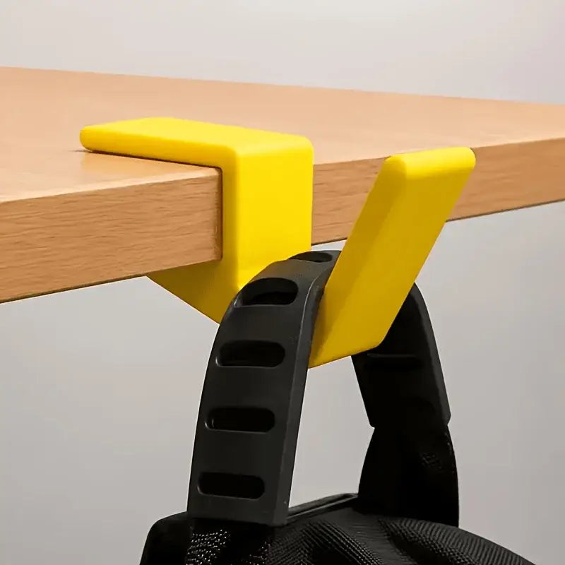 bright yellow plastic desk clamp holding a black backpack strap as a desk edge bag hook for convenient backpack storage