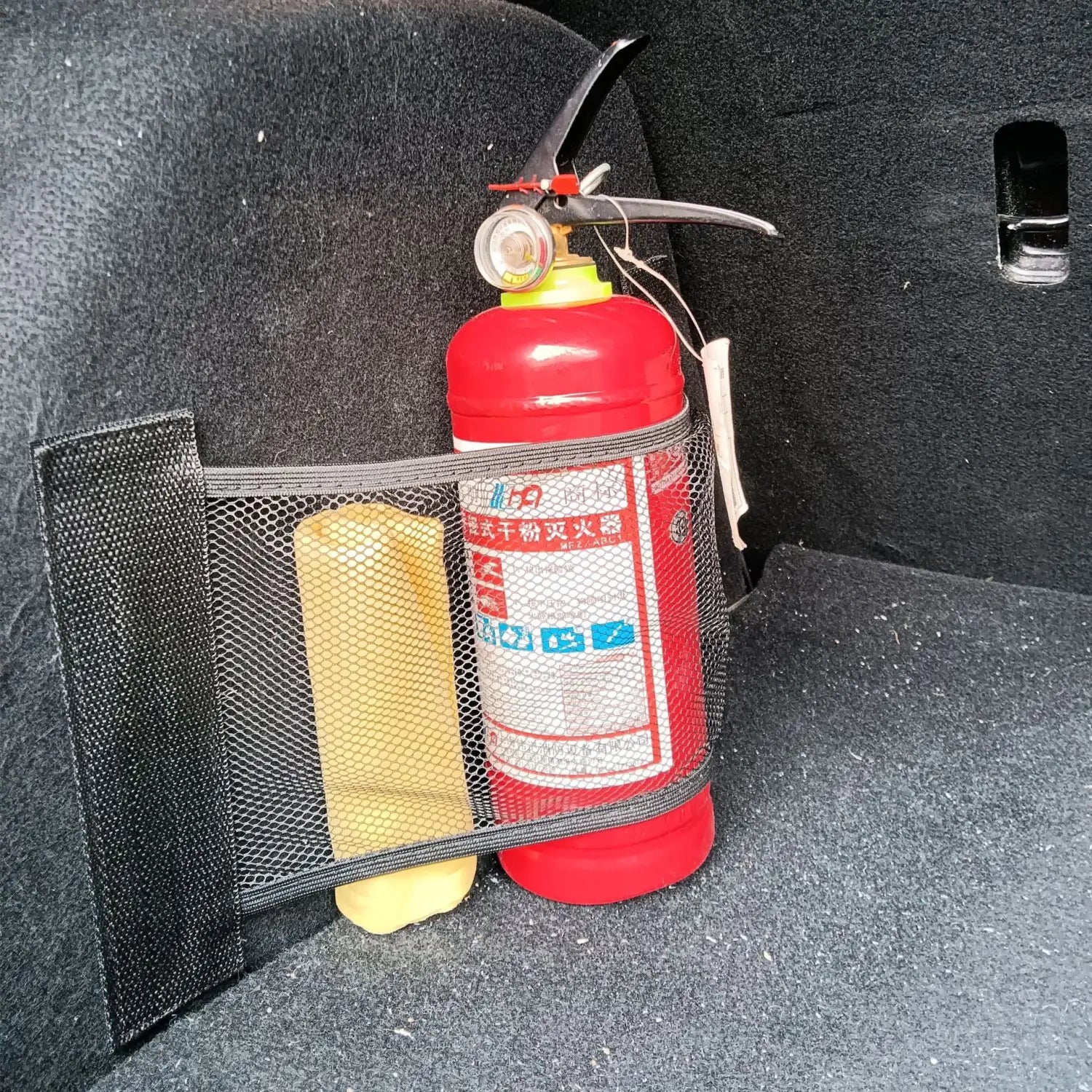 Roter Feuerlöscher mit gelbem Kopf und schwarzem Griff, gesichert in einem schwarzen elastischen Aufbewahrungsnetz für die Kofferraumorganisation.