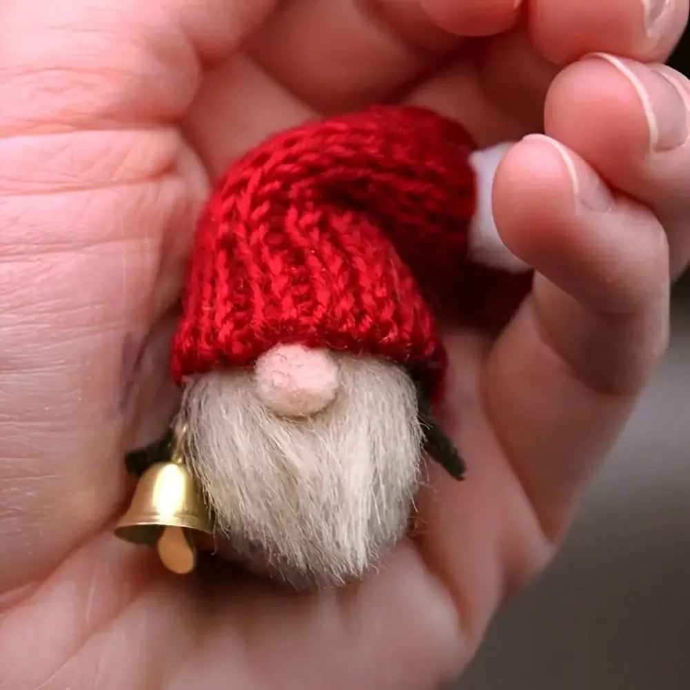 Eine winzige, gestrickte rote Mütze mit flauschigem weißem Bart und rosiger Nase, die einen kleinen Mini-Glockenanhänger aus goldenem Material für Bastelarbeiten DIY hält.