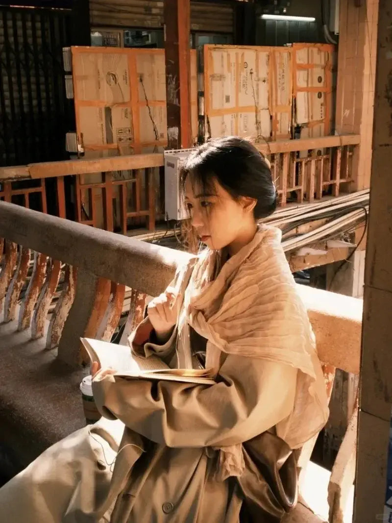 Una mujer sentada y leyendo lleva una bufanda de algodón lino color beige para mujer de color sólido y chal transpirable, combinada con un abrigo tipo trench a juego.