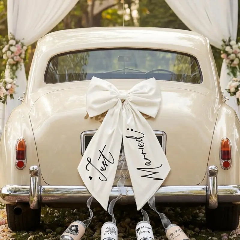 Une voiture ancienne de couleur crème décorée d’un grand nœud de voiture « just married » en satin blanc, avec une écriture noire cursive élégante, pour la décoration de mariage.