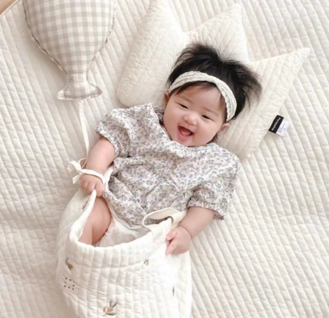Ein entzückendes Baby, das neben einem gepolsterten Babywindel-Aufbewahrungskörbchen sitzt, in einem weichen hellgrauen Kleid mit floralem Muster, kurzen Ärmeln und gerafftem Halsausschnitt.