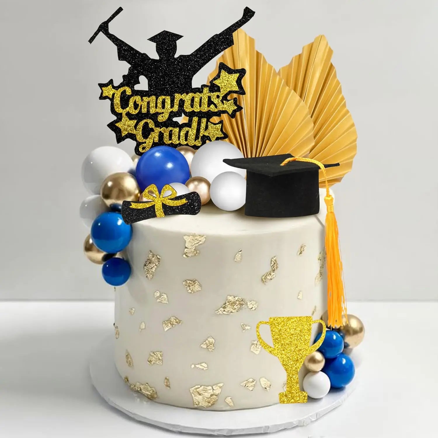 A white graduation cake adorned with gold leaf and a glittery congrats grad topper is accompanied by blue and gold balloons and a black graduation cap with a yellow tassel as part of a graduation cake topper set for 2026 senior party.