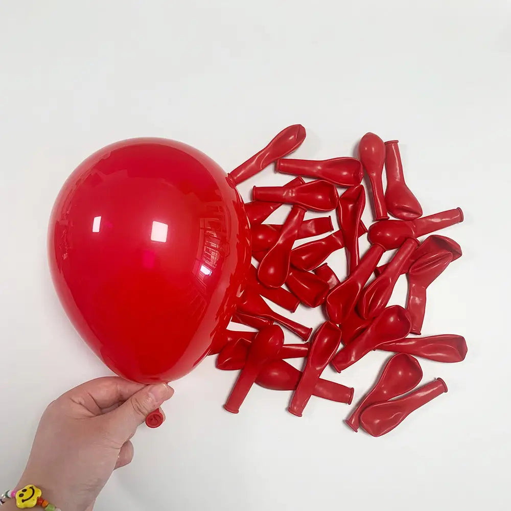 Eine Hand, die einen einzelnen aufgeblasenen roten Latexluftballon hält, mit zahlreichen entleerten roten Latexluftballons, die in der Nähe verstreut sind, in einer Partydekoration „Rot Schwarz Weiß Ballon-Set“.