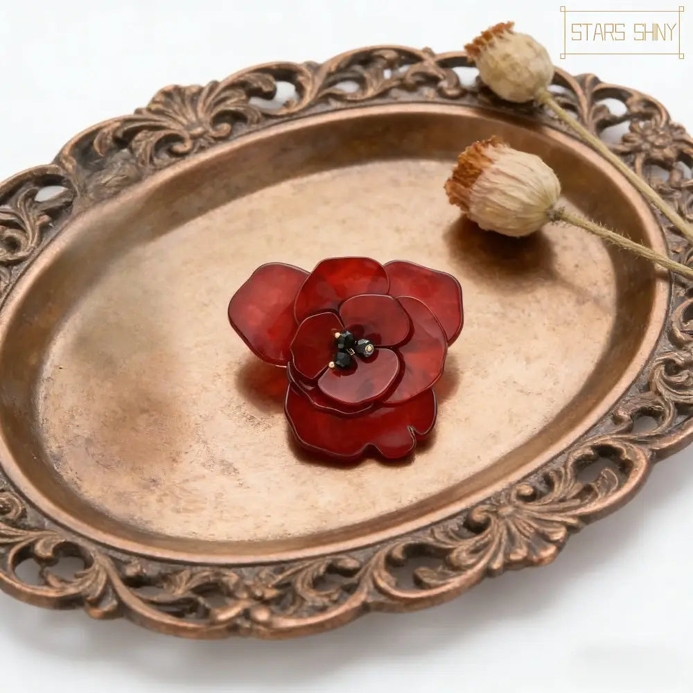 Une broche en verre rouge laquée en forme de fleur, avec un centre noir et de minuscules accents métalliques, dans le style d’une épingle coquelicot en émail de fleur rouge.