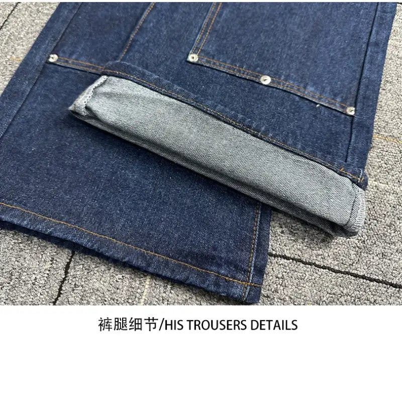 Jeans bruts en denim à jambe large à rivets pour hommes en bleu foncé, avec des coutures renforcées et des fermetures boutonnées à finition argentée, les revers retroussés laissant apparaître un tissu intérieur gris clair et des coutures renforcées.