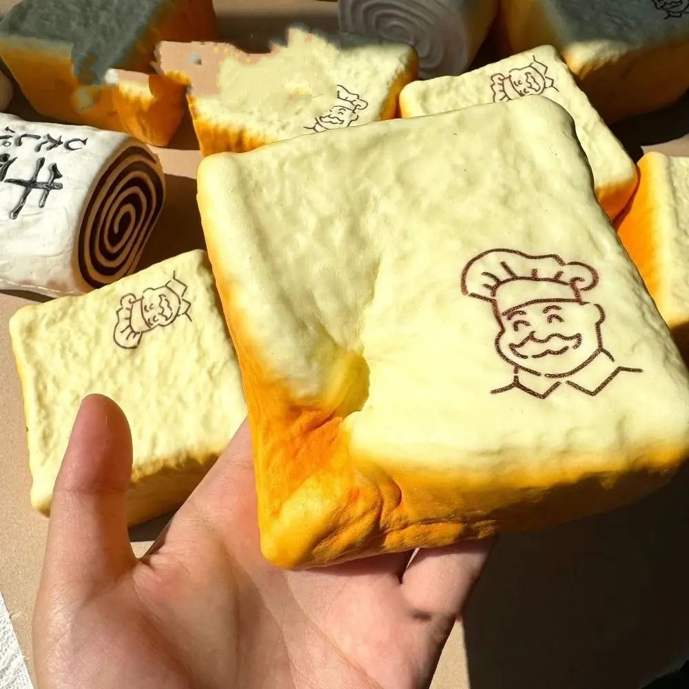 a pale yellow squishy toy shaped like a bread slice with a golden-brown crust and a black line drawing of a smiling chef on the front, designed as a sensory fidget toy suitable for a universal age range