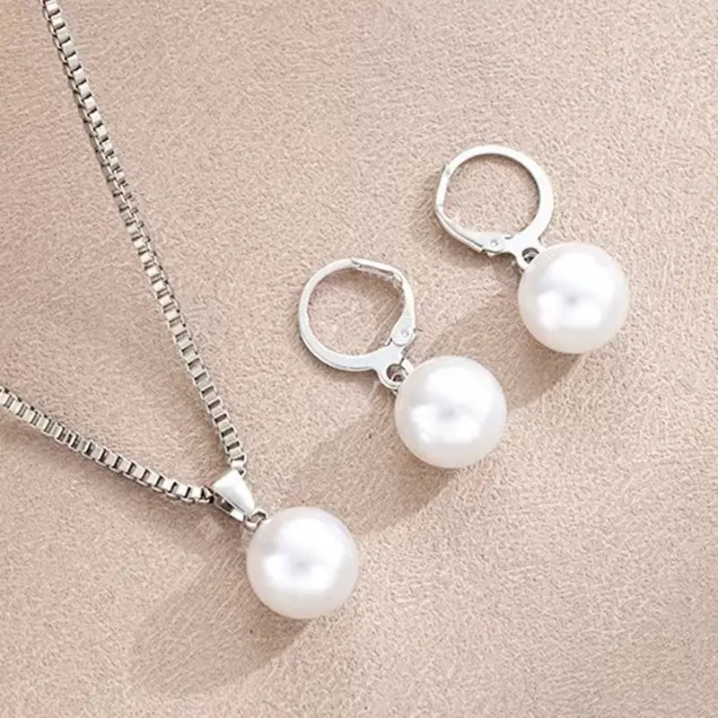 Un ensemble de bijoux en tons argentés, collier à pendentif et boucles d’oreilles créoles assorties, chacun arborant une seule perle ronde blanche.