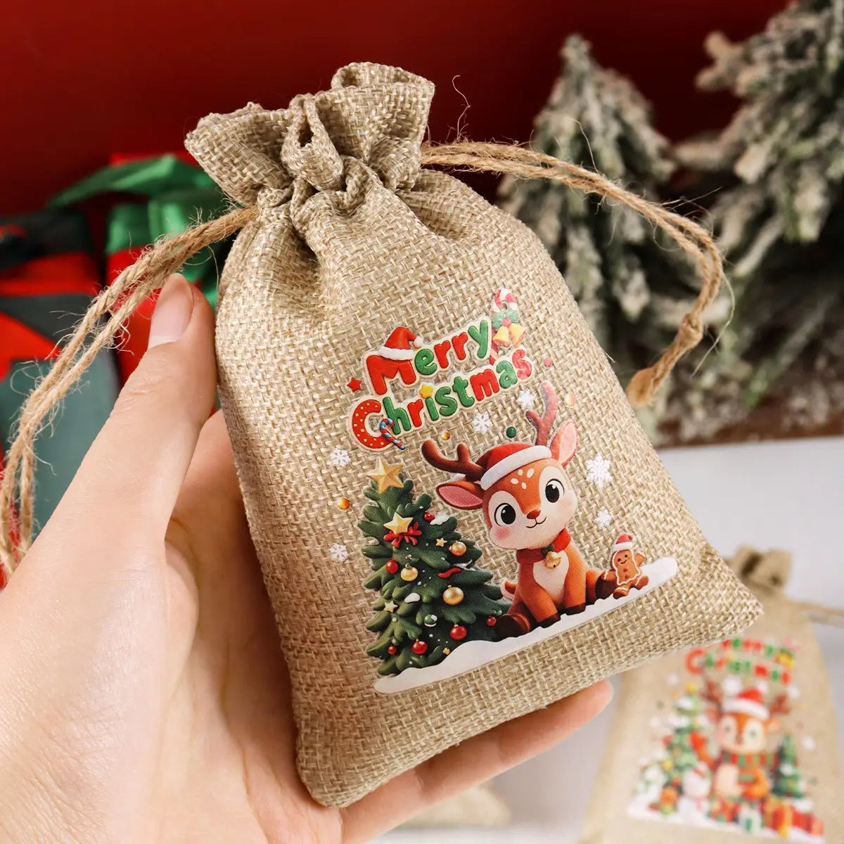 Sachet cadeau en toile de jute beige à cordon coulissant provenant du lot de six sachets cadeaux en toile de jute pour Noël, avec un motif festif « Joyeux Noël » et un renne de style dessin animé, 圣诞树, ainsi qu’un homme en pain d’épices.