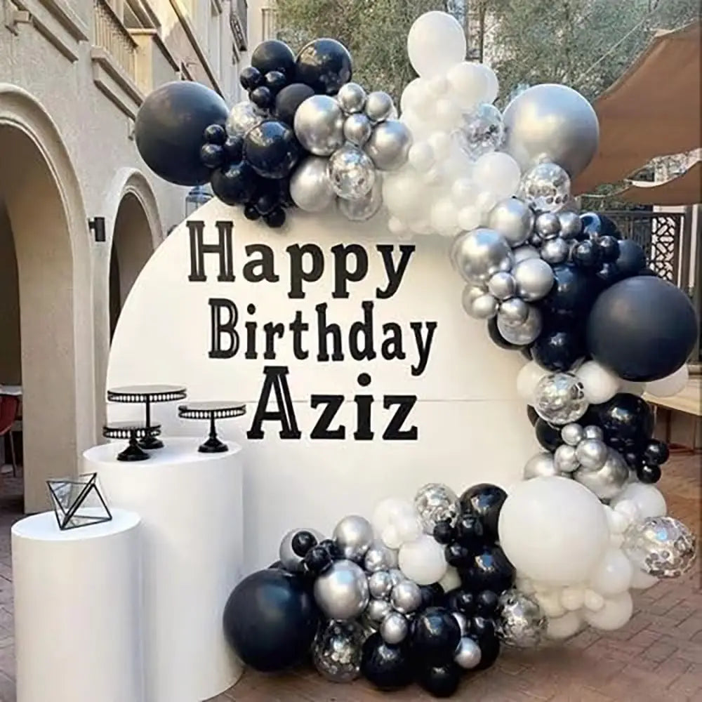 Ein festliches Ballonbogen-Set aus Schwarz, Weiß und Silber für Partys mit einem Happy-Birthday-Aziz-Schild.