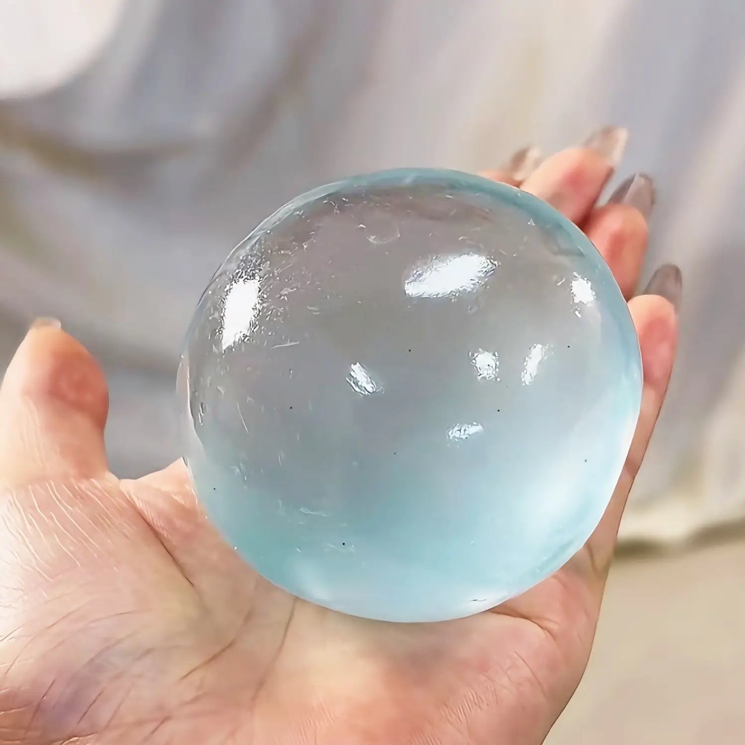 Une balle anti-stress translucide bleu clair de type gel, avec une surface lisse et brillante et des reflets internes subtils, tenue dans une main, pour une utilisation comme jouet de compression