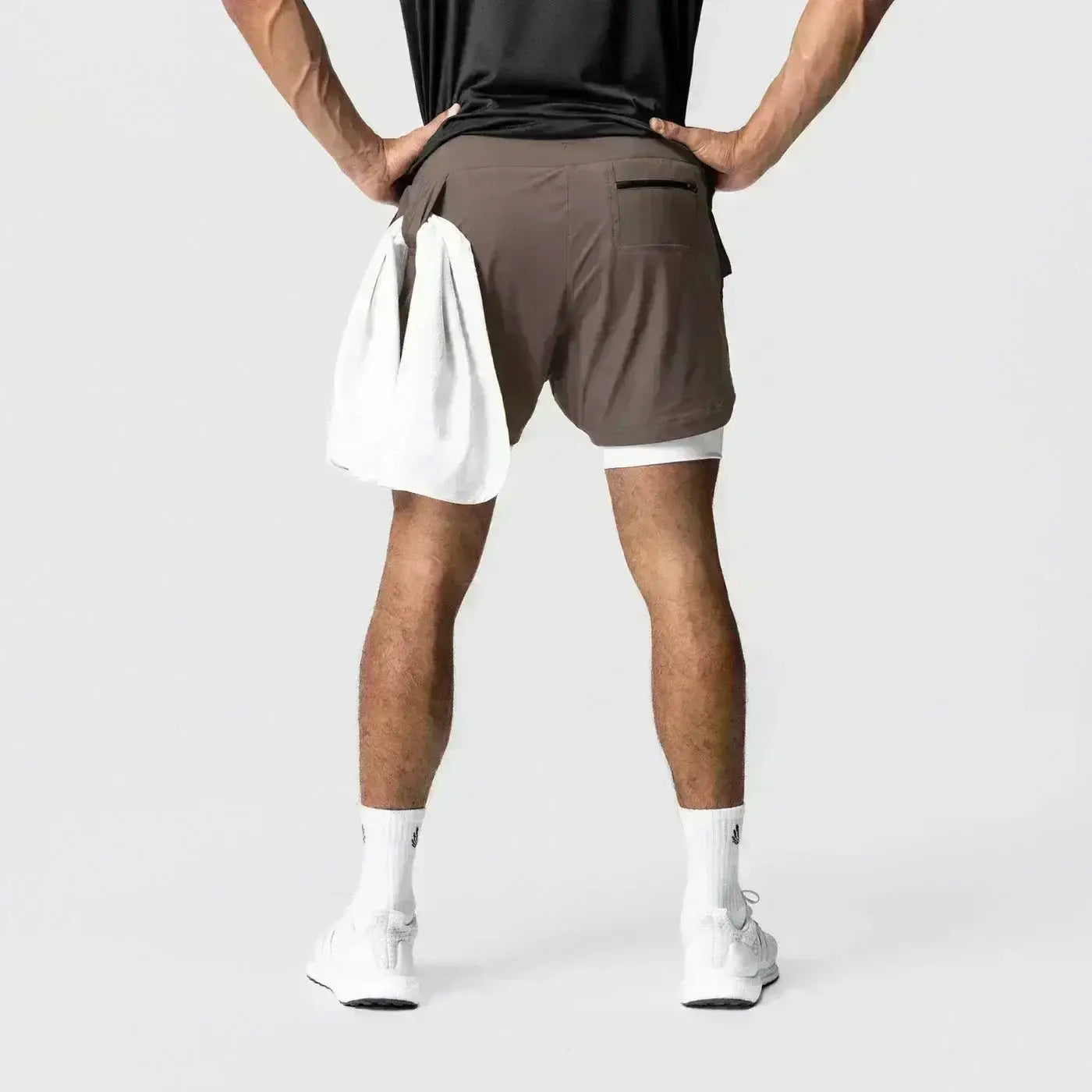 Hombres, pantalones cortos atléticos 2 en 1 para gimnasio color marrón con capa interior blanca, bolsillos laterales y una trabilla para la toalla que ofrece soporte, mostrados desde el lado posterior, usados por una persona que está de espaldas una a la otra.