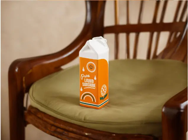 Vase en céramique en forme de brique de jus d’orange, orange vif, avec un dessus blanc et un décor de soleil ludique, étiqueté « pure liquid sunshine ».
