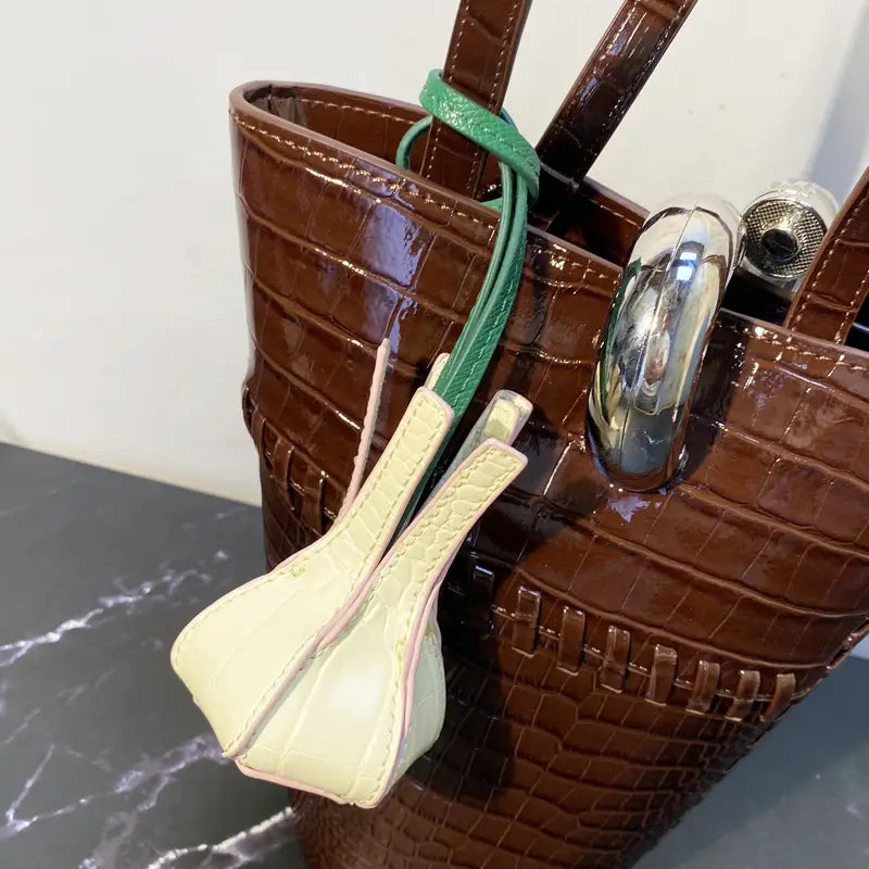 Un tote bag en cuir verni façon crocodile, brun, présentant des charms pour sac à main en cuir végétal vert et crème, conçus comme ornements de sac à main de créateur.