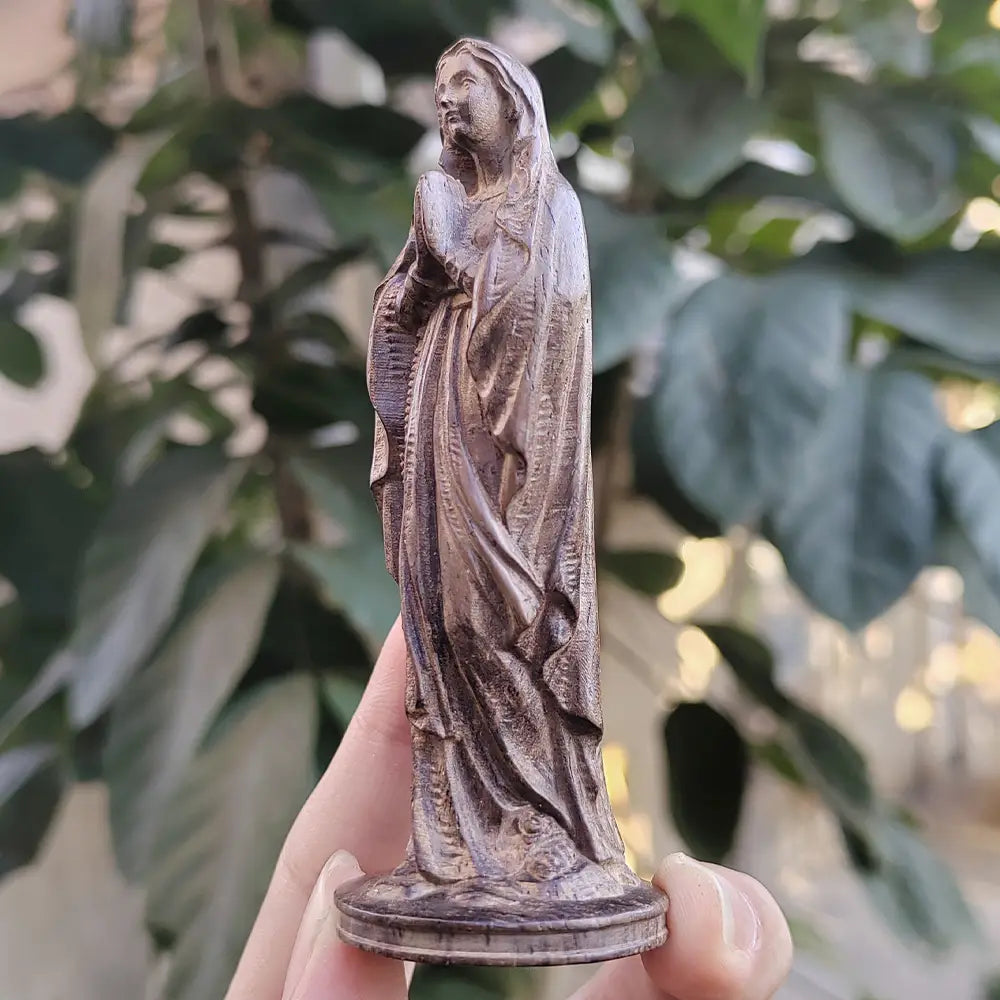 Statuette détaillée de couleur bronze représentant une Vierge Marie vêtue d’une robe, avec de longs cheveux et les mains jointes en prière, se tenant sur un socle circulaire sculpté à la main dans du bois.