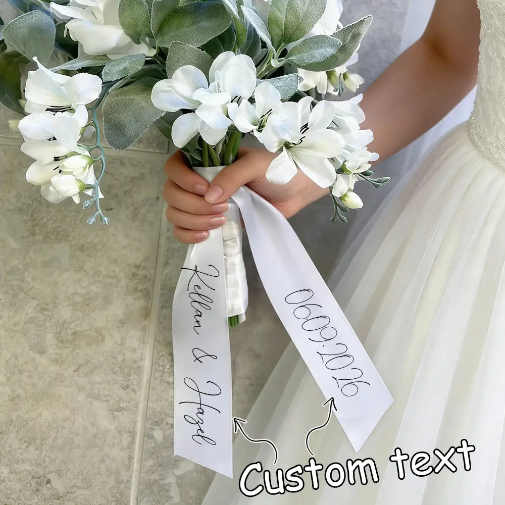 Un délicat bouquet de mariage artificiel blanc et argenté, comportant des rubans de texte personnalisés avec des noms propres et une date.