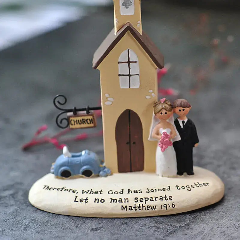 Figura decorativa de boda en resina color beige y marrón que muestra a una novia con vestido blanco sosteniendo flores rosadas, un novio con atuendo negro, un coche azul pequeño y una base grabada con la frase «por lo tanto, lo que Dios ha unido, que no lo separe el hombre», diseñada como regalo de aniversario.