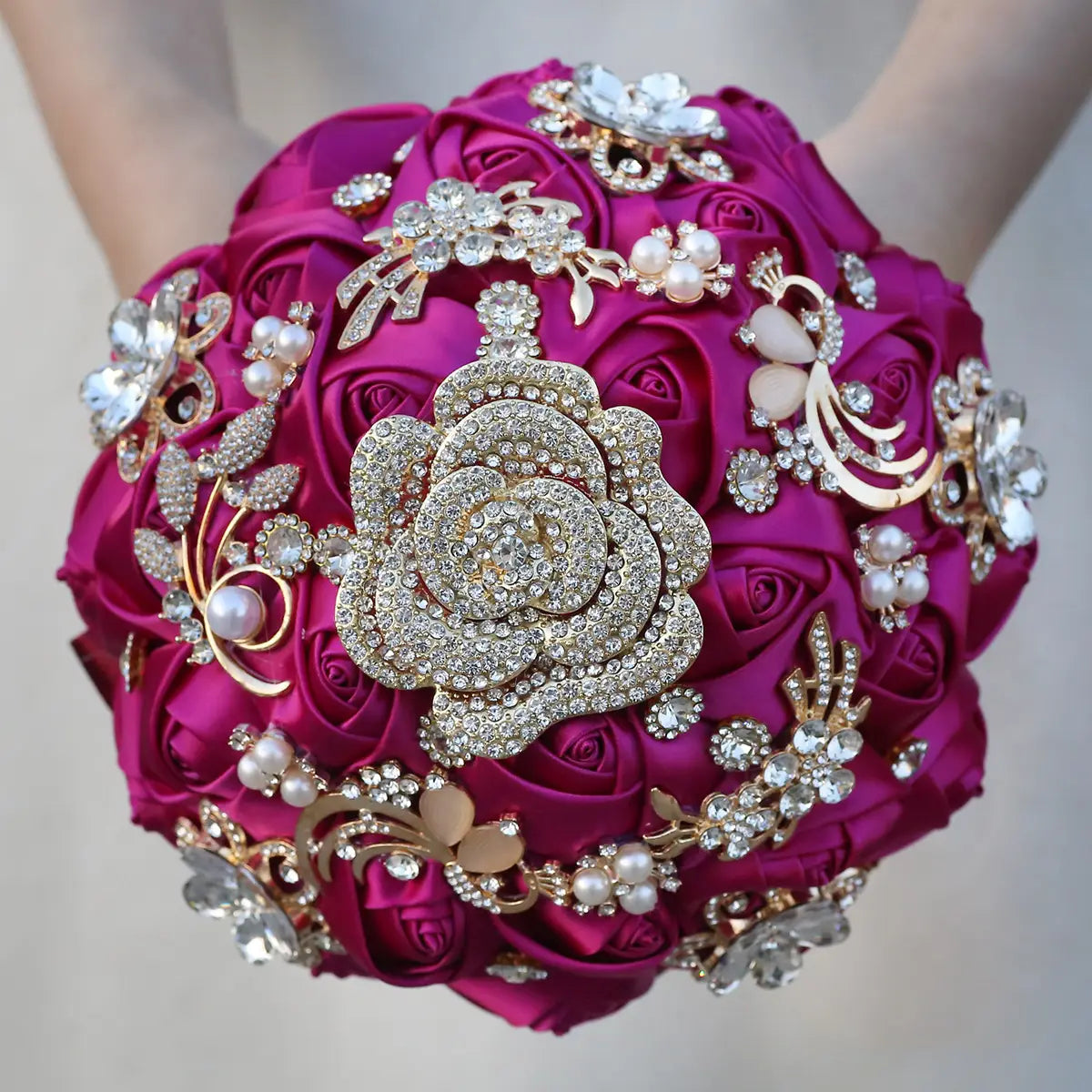 Un bouquet de mariée vibrant en satin fuchsia orné de broches finement ouvragées aux tons dorés, de strass étincelants et d’accents de perles.