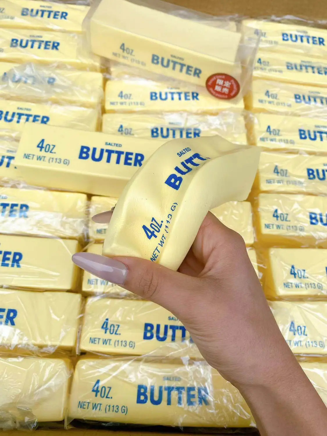 Una mano sostiene un único juguete antiestrés blando con forma de barra de mantequilla, de color amarillo pálido, que asciende lentamente, envuelto en plástico transparente con letras azules y marcas de peso.