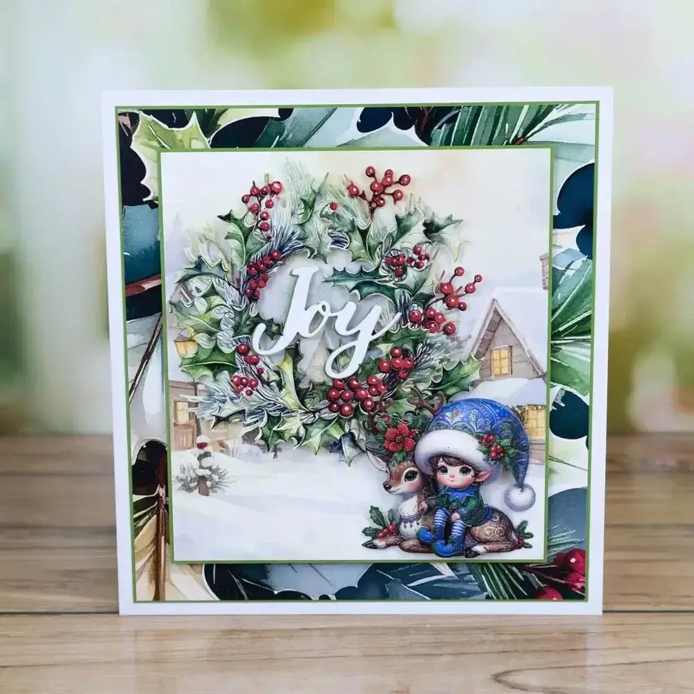 a Christmas metal cutting die and stamp set featuring a festive card design with a detailed holly wreath with red berries, the word joy in white script, and two illustrated characters including a child in a blue hat and a deer against a snowy village scene.